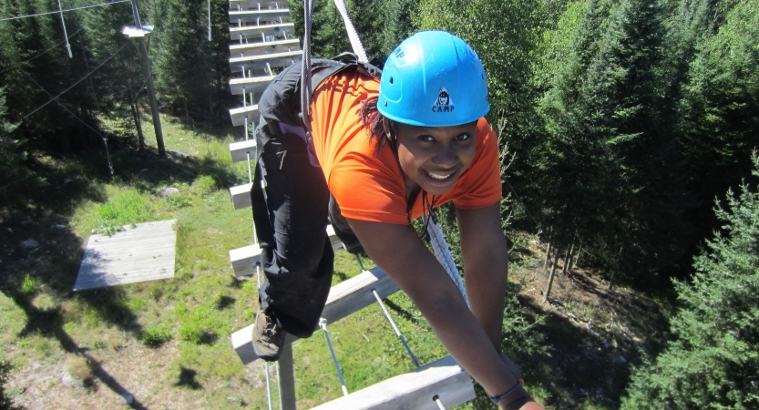 A person wearing safety gear and secured by ropes smile as they balance on an obstacle on a high ropes course. 